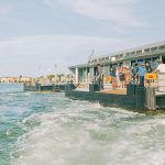 Turistas chegam ao terminal de balsas do Lido, em Veneza, com a paisagem e a arquitetura da região ao fundo.