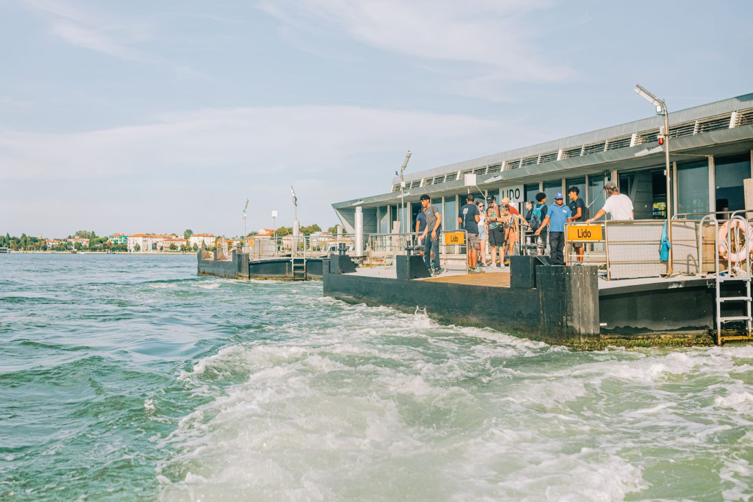 Turistas chegam ao terminal de balsas do Lido, em Veneza, com a paisagem e a arquitetura da região ao fundo.