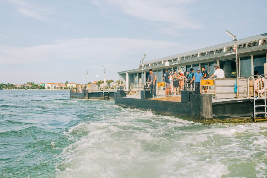 Turistas chegam ao terminal de balsas do Lido, em Veneza, com a paisagem e a arquitetura da região ao fundo.