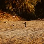 Duas crianças soltam pipa em um campo de terra cercado por vegetação.