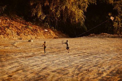 Duas crianças soltam pipa em um campo de terra cercado por vegetação.