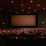 Sala de cinema vazia, com fileiras de poltronas vermelhas diante de uma tela iluminada.