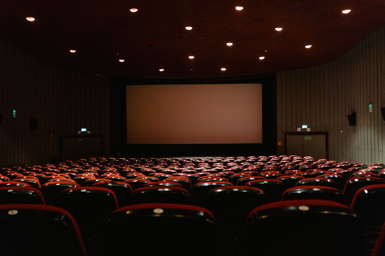 Sala de cinema vazia, com fileiras de poltronas vermelhas diante de uma tela iluminada.