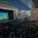 Público reunido à noite para assistir a uma exibição de filme ao ar livre sob uma ponte.
