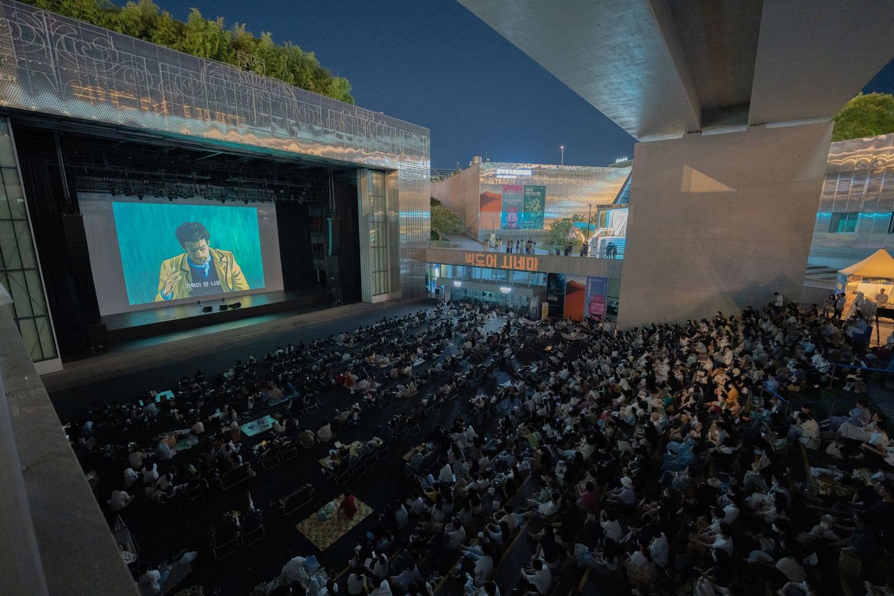 Público reunido à noite para assistir a uma exibição de filme ao ar livre sob uma ponte.