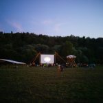 Pessoas reunidas em um campo ao entardecer para assistir a uma sessão de cinema ao ar livre.