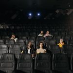 Plateia diversificada parcialmente sentada em uma sala de cinema pouco iluminada, diante da tela.
