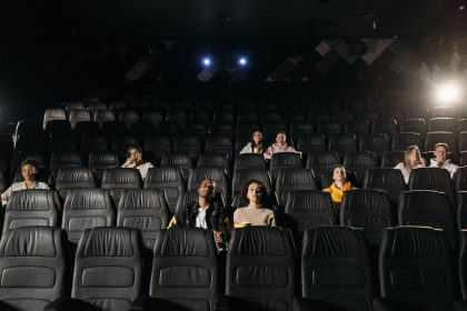 Plateia diversificada parcialmente sentada em uma sala de cinema pouco iluminada, diante da tela.