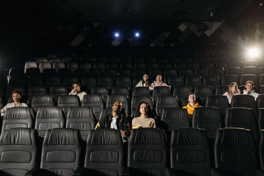 Plateia diversificada parcialmente sentada em uma sala de cinema pouco iluminada, diante da tela.
