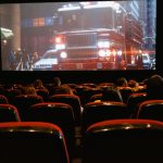 Público sentado em uma sala de cinema enquanto assiste a um filme projetado em uma grande tela.